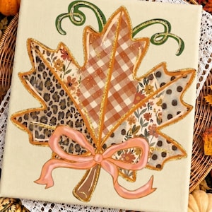 May include: A square canvas featuring a large maple leaf illustration divided into sections with leopard print, brown and white gingham, floral patterns, and polka dots. The leaf has gold glitter outlines, green curly stems, and a peach ribbon bow.