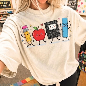 May include: A cream-colored t-shirt featuring a graphic print of four anthropomorphic school supplies: a yellow pencil, a red apple, a black composition notebook, and a blue ruler, all with smiling faces, arms, and legs, surrounded by small stars and hearts.