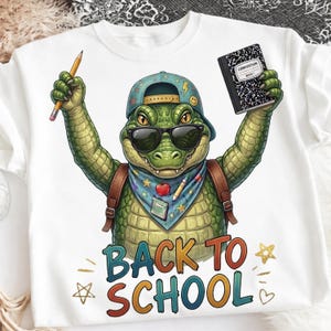 Puede incluir: Una camiseta blanca con el gráfico de un caimán verde que lleva una gorra azul, gafas de sol y un pañuelo. El caimán sostiene un lápiz amarillo y un cuaderno de composición en blanco y negro. El texto BACK TO SCHOOL aparece impreso debajo.