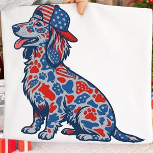 May include: A white fabric square featuring a graphic illustration of a dog wearing a baseball cap. The dog is patterned with red, white, and blue shapes, including stars, stripes, and paw prints, reflecting a patriotic theme in its design.