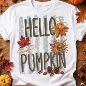 May include: A white t-shirt featuring the text HELLO PUMPKIN in brown letters. The design includes a light blue striped background with white polka dots, decorated with autumn elements like a pumpkin, maple leaves, wheat, and pinecones.