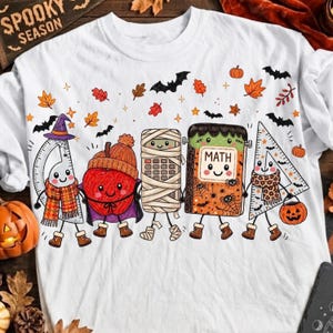 Op de afbeelding: Een wit t-shirt met een afbeelding van vijf antropomorfe schoolspullen in Halloween-kostuums, waaronder een gradenboog, een appel, een rekenmachine, een wiskundeboek en een geodriehoek, omringd door vleermuizen, bladeren en pompoenen.