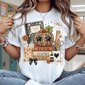 May include: A white t-shirt featuring a graphic print of a leopard-print pumpkin wearing star-shaped sunglasses, stacked books labeled TEACH INSPIRE LOVE, a notebook labeled teacher notes, a coffee cup, a glue bottle, scissors, and bows.