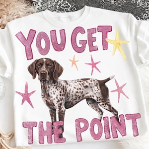 Op de afbeelding: Een wit sweatshirt met ronde hals en een bruin-wit gevlekte hond in het midden. De tekst YOU GET THE POINT staat in grote roze letters boven en onder de hond, omringd door kleine gele, oranje en roze zeesterren.