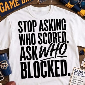 May include: A white short-sleeved t-shirt featuring bold black text that reads: STOP ASKING WHO SCORED. ASK WHO BLOCKED. The lettering is arranged in a stacked format with a mix of standard sans-serif and stylised brush-stroke fonts for emphasis.