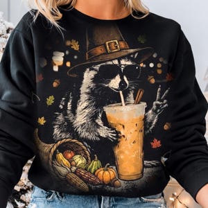 May include: A black long-sleeve sweatshirt featuring a graphic of a raccoon wearing a brown pilgrim hat and black sunglasses. The raccoon holds an iced coffee with a cinnamon stick and makes a peace sign. A cornucopia with autumn vegetables sits below.