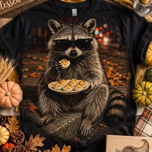 Puede incluir: Una camiseta negra con un estampado gráfico de un mapache con gafas de sol negras. El mapache está sentado al aire libre, sosteniendo un huevo relleno en una mano y equilibrando un plato de huevos rellenos en la otra sobre un fondo otoñal.