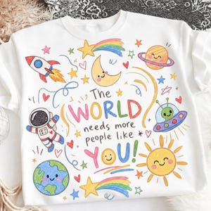 May include: A white sweatshirt featuring a colourful crayon-style illustration of space elements including a rocket, planet, moon, astronaut, Earth, UFO, and sun. The text reads, The WORLD needs more people like YOU! surrounded by small stars and hearts.