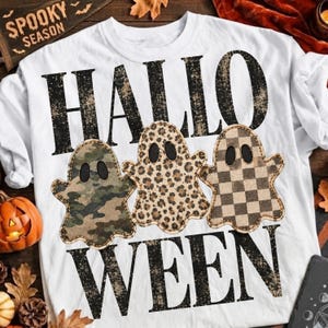 May include: A white t-shirt featuring the black distressed text "HALLO" above three ghost shapes and "WEEN" below. The ghosts have black eyes and feature different patterns: a green camouflage print, a brown leopard print, and a brown checkered print.