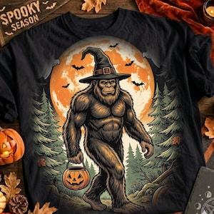 Może przedstawiać: Czarny t-shirt z grafiką przedstawiającą brązowego Wielką Stopę w kapeluszu czarownicy, trzymającego wiaderko w kształcie dyni. W tle znajduje się duży pomarańczowy księżyc w pełni, drzewa iglaste oraz latające nietoperze na środku koszulki.