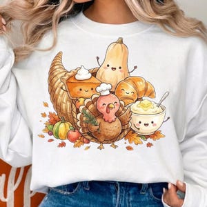 May include: A white long-sleeve sweatshirt featuring a graphic print of a cornucopia filled with a smiling turkey wearing a chef hat, a slice of pumpkin pie, a butternut squash, a croissant, and a bowl of mashed potatoes with butter.