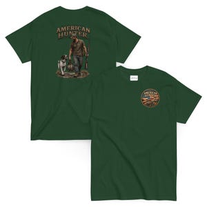 Könnte beinhalten: Ein dunkelgrünes Kurzarm-T-Shirt mit einem kreisförmigen Logo auf der Brust, das ein Gewehr und eine amerikanische Flagge zeigt. Auf der Rückseite befindet sich eine große Abbildung eines Jägers mit Hund und der Schriftzug AMERICAN HUNTER.