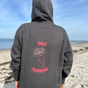 Könnte beinhalten: Ein anthrazitfarbener Kapuzenpullover mit einer Grafik auf der Rückseite. Das Design enthält den roten Schriftzug "OVER" und "THINKING" sowie eine stilisierte Strichzeichnung eines Gesichts und eine verworrene Gehirnform.