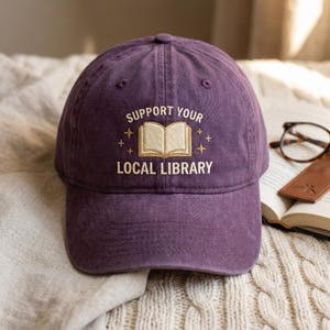 May include: A purple baseball cap with the embroidered text "SUPPORT YOUR LOCAL LIBRARY" and a book graphic. The hat is made of a soft material and has a curved brim. A pair of glasses and a book are in the background.