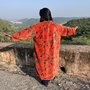 Pode incluir: Um casaco comprido de veludo laranja até ao joelho, de manga comprida, com um estampado vibrante de flores e pássaros. A peça apresenta uma silhueta larga e aberta à frente, com mangas largas e um acabamento de tecido macio e texturado.