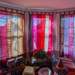 May include: A set of three window curtains featuring vertical panels of sheer fabric in shades of red, pink, and brown with various intricate patterns. The curtains are hung from a dark rod using fabric loops and cover a bay window arrangement.