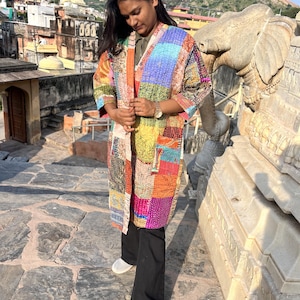 May include: A long-sleeved, knee-length open jacket featuring a vibrant patchwork design made from various colourful fabrics with intricate kantha stitching. The garment includes a solid-coloured collar and two front pockets, showcasing a mix of patterns.