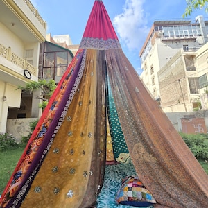 May include: A conical hanging canopy tent constructed from multiple panels of sheer, multicoloured fabric featuring various patterns, floral prints, and metallic embellishments, with a solid pink top section and a patterned teal floor mat.
