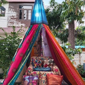 May include: A conical fabric tent featuring panels of vibrant, multicoloured patterned textiles in shades of pink, orange, blue, and green. Inside the tent sits an upholstered armchair covered in floral fabric, accented with several decorative throw pillows.
