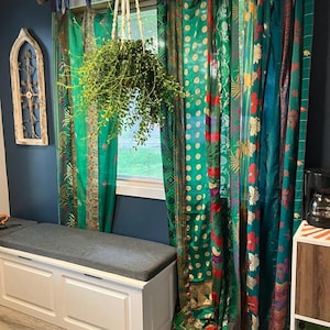 Pode incluir: Cortinas de comprimento total com um patchwork de painéis de tecido verde, azul-petróleo e vermelho com vários padrões florais, geométricos e de pavão. As cortinas pendem em frente a uma janela, ocultando parcialmente uma planta suspensa.