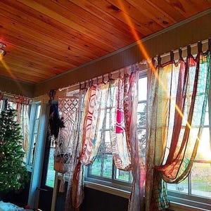 May include: A cozy interior with a wooden ceiling and patterned curtains. Sunlight streams through the windows, illuminating a Christmas tree and decorative macrame. The curtains are in shades of red, orange, and green.