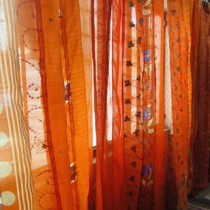 Peut inclure: Une série de rideaux orange transparents suspendus à une tringle, présentant des motifs variés incluant des rayures verticales blanches, de grands pois blancs et une délicate broderie florale noire avec des accents de petites fleurs bleues.