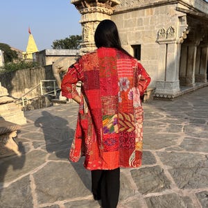 Könnte beinhalten: Eine langärmelige Jacke mit einem Patchwork-Design aus verschiedenen rotfarbenen Stoffen mit durchgehenden, komplexen weißen Nähten. Das Kleidungsstück zeigt eine Mischung aus floralen und geometrischen Mustern in Orange, Pink und Dunkelrot.