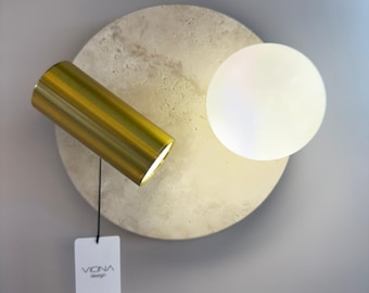 May include: A modern wall sconce featuring a cylindrical gold-toned light fixture angled towards a round, off-white, textured concrete base. A white, spherical light is adjacent to the base. A tag with the text "viona design" hangs below.