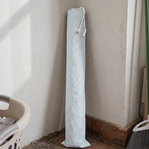 May include: A tall, cylindrical fabric storage bag in a light blue colour featuring a repeating pattern of small pink and yellow floral motifs. The bag is secured at the top with a white braided drawstring cord that has two knotted tassel ends.