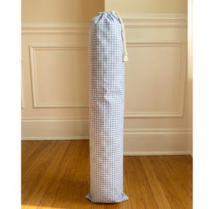 May include: A tall, cylindrical fabric storage bag featuring a light blue and white gingham check pattern. The bag is secured at the top with a white drawstring cord, creating a gathered closure. The structure is upright and elongated in shape.
