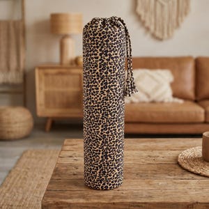 Pode incluir: Uma bolsa cilíndrica de tecido para tapete de ioga com um padrão de leopardo em preto e bege. A bolsa possui um fecho de cordão na parte superior com duas borlas de tecido a condizer penduradas no cordão para transportar o tapete enrolado.
