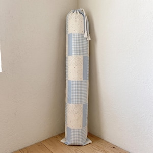May include: A tall, cylindrical fabric storage bag featuring a patchwork design of light blue gingham checks and a cream-coloured fabric with small, scattered brown dots. The bag is cinched at the top with a white drawstring cord and tassels.