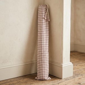 May include: A tall, cylindrical fabric storage bag featuring a mauve and white gingham check pattern. The bag is cinched at the top with a matching drawstring cord that includes two small tassels at the ends, providing a secure closure.