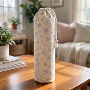Pode incluir: Um saco cilíndrico de tecido em creme com um padrão repetitivo de linhas azuis com ursinhos de peluche, laços, cerejas e pequenos motivos florais. O saco é fechado no topo com um cordão entrançado grosso de cor creme e uma borla.
