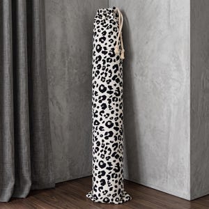 Pode incluir: Um saco de armazenamento cilíndrico de tecido com um padrão de leopardo em preto e branco. O saco inclui um fecho de cordão no topo com cordas grossas de cor creme, concebido para o armazenamento vertical de objetos longos e finos.