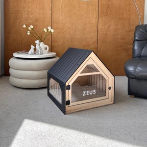 Puede incluir: Una caseta para perros de madera con techo negro y paredes de acrílico transparente. La caseta tiene un marco de madera de color claro y una cama en el interior. La palabra "ZEUS" está impresa en la parte delantera. Una huella de pata está en la puerta.