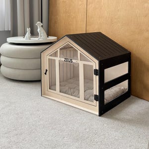 以下が含まれることがあります： 黒い切妻屋根と明るい色のフレームを持つ木製のペットハウス。前面には透明なドアがあり、骨の形をした切り抜きに「ZEUS」という名前が表示されています。内部の床にはベージュのキルティングクッションが敷かれています。