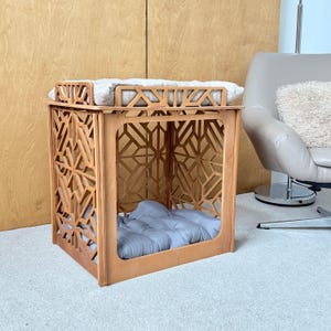 May include: A light brown wooden pet crate with an intricate geometric lattice pattern on the sides and top. The structure features a flat top surface with a light-colored tufted cushion and a matching grey tufted cushion placed inside the base.