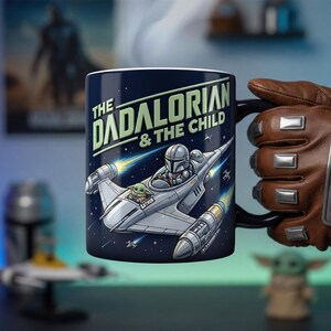 以下が含まれることがあります： 「The Dadalorian & The Child」の文字と、マンダロリアンとグローグーが描かれた宇宙船の漫画のイラストが特徴の、濃い青色のセラミックマグカップ。マグカップは黒いハンドルが付いており、茶色の手袋をはめた人が持っています。