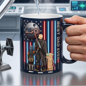 May include: A navy blue ceramic mug with a black handle features a design with the text "BEST DAD EVER." The mug showcases a Star Wars theme with a Death Star, the American flag, and characters. The names "JENNY," "DAVID," and "ANNA" are printed at the bottom.