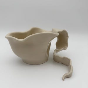 May include: A cream-coloured ceramic jug with a smooth, glossy finish. The vessel features a wide, undulating rim and a long, ribbon-like handle that extends outwards and curves downwards, creating a distinct, flowing silhouette.