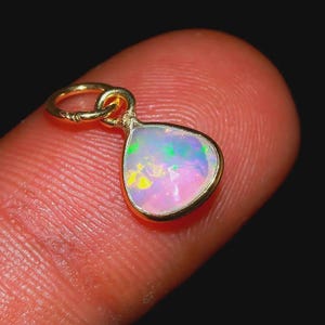 Può includere: Un ciondolo in opale a forma di goccia incastonato in una montatura in metallo dorato con un piccolo anello circolare sulla parte superiore. L'opale presenta una base bianca traslucida con riflessi iridescenti verdi, blu e rosa.