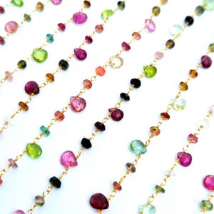 Può includere: Diverse catene in metallo dorato con piccole pietre preziose sfaccettate a forma di goccia in vari colori, tra cui rosa, verde, giallo, nero e trasparente. Le pietre sono distanziate uniformemente lungo le sottili maglie metalliche.
