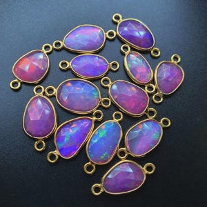 May include: A collection of faceted, oval and teardrop-shaped purple opal gemstones with gold-colored settings. Each stone has a unique play of color, with flashes of blue, green, and red. The gemstones are arranged on a dark surface.