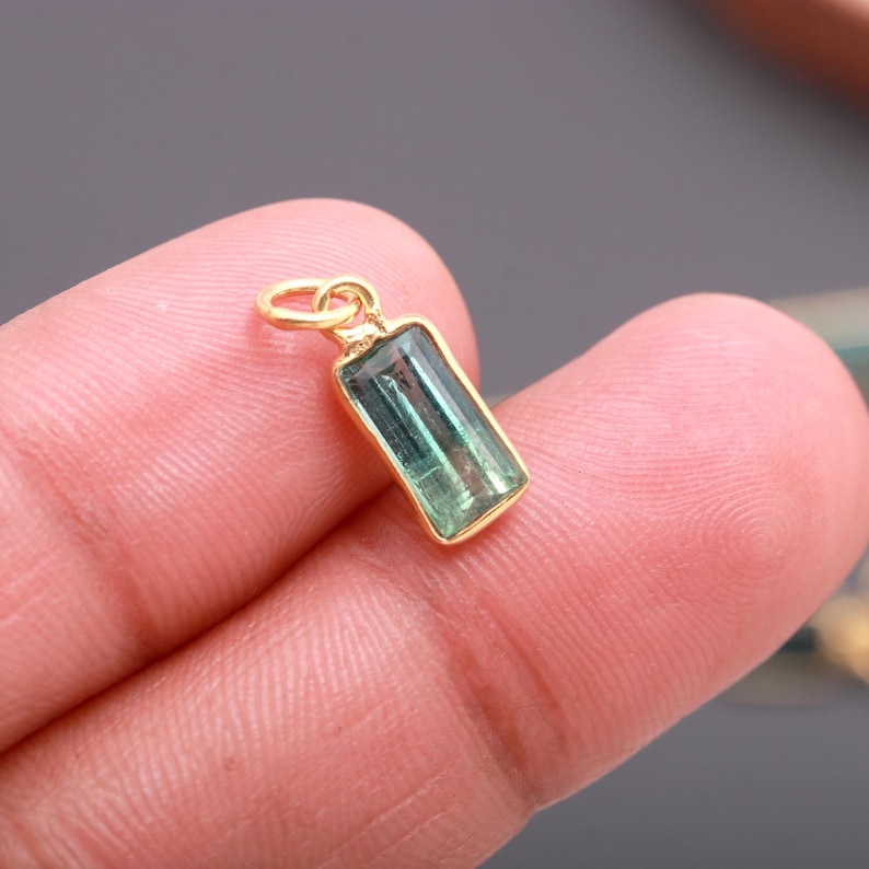 K&ouml;nnte beinhalten: Ein rechteckiger gr&uuml;ner Turmalin-Edelsteinanh&auml;nger in einer goldfarbenen Metallfassung mit einem kleinen runden Verbindungsring an der Oberseite. Der Edelstein hat eine durchscheinende, facettierte Oberfl&auml;che mit sichtbaren Einschl&uuml;ssen.