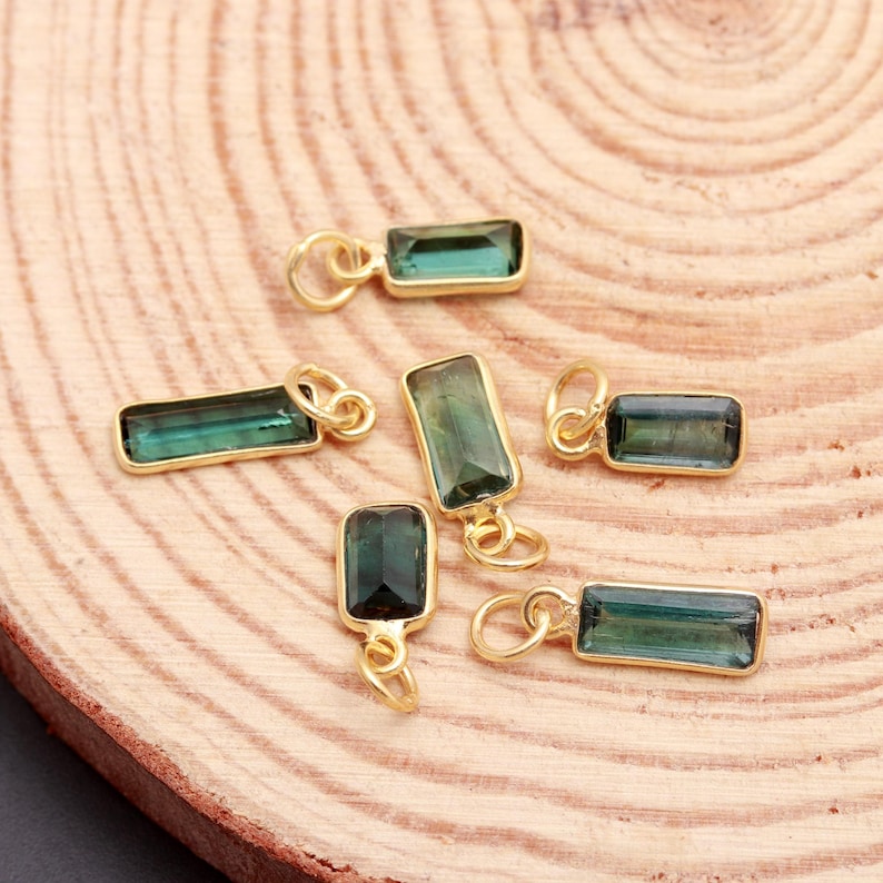 K&ouml;nnte beinhalten: Eine Ansammlung von sechs rechteckigen Anh&auml;ngern, die jeweils einen facettierten dunkelgr&uuml;nen Edelstein in einer goldfarbenen Metallfassung zeigen, mit einem kleinen runden Biegering an der Oberseite zum Aufh&auml;ngen, verstreut angeordnet.