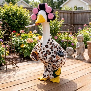 May include: A white goose figurine wearing a leopard print robe and pink hair rollers. The goose has a yellow beak holding a cigarette, a round orange object in one wing, and a black coffee cup in the other wing. The figure stands on yellow feet.