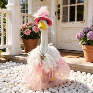 Puede incluir: Una estatua de ganso blanco que lleva un sombrero rosa con una pluma, una boa de plumas blancas y una falda de tul rosa con pequeños detalles de estrellas doradas. El ganso tiene un pico amarillo y está en posición vertical.