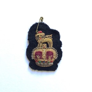 May include: A gold embroidered crown with a red velvet center and a gold lion on top. The crown is on a black felt backing with a safety pin attached.