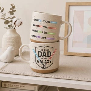 May include: Two beige ceramic mugs stacked on top of each other. The top mug has the names Ethan, Mia, Leo, and Ava with colored lightsaber designs. The bottom mug reads "THE BEST DAD IN THE GALAXY" with lightsaber graphics.